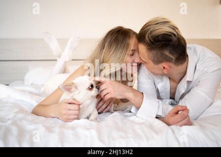 Una coppia coccola un cane seduto sul letto di casa. Un uomo e una donna si siedono con il loro cane sul letto e giocano con esso. pet un chihuahua. Foto Stock
