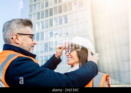 L'ingegnere maschile regola il casco per l'ingegnere femminile prima di entrare nel business center - due colleghi ingegneri che indossano giubbotti da lavoro arancioni Foto Stock