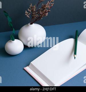 Taccuino e matita vuoti con vaso di fiori asciutti sulla scrivania. Sfondo blu scuro parete. Spazio di lavoro Foto Stock