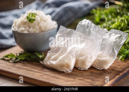 Riso crudo in sacchetti di plastica su sfondo di legno e riso cotto in ciotola. Foto Stock