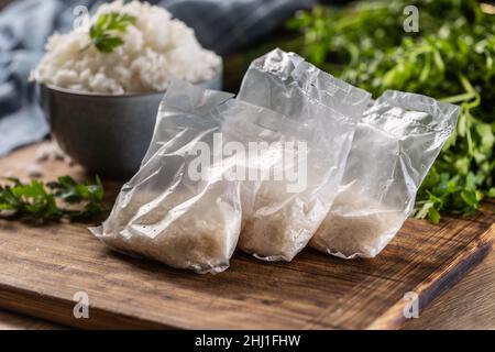 Riso crudo in sacchetti di plastica su sfondo di legno e riso cotto in ciotola. Foto Stock