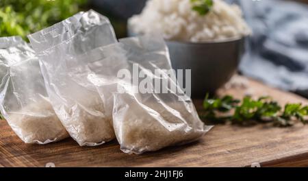 Riso crudo in sacchetti di plastica su sfondo di legno e riso cotto in ciotola. Foto Stock