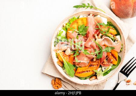 Ciotola per insalate con pesca alla griglia, jamon, formaggio feta, noci e rucola su sfondo bianco del tavolo, vista dall'alto, spazio per copie Foto Stock