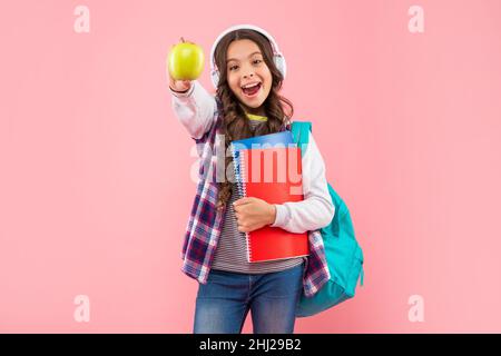 stupito bambino ascoltare musica in auricolari con borsa scuola e libro di esercizi tenere mela, scuola. Foto Stock