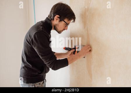 Uomo strappare a mano la vecchia carta da parati con raschietto dal muro. Lavori di ristrutturazione della casa. Foto Stock