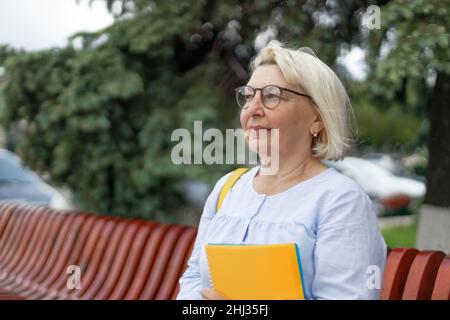 Donna insegnante di 50 anni su una panca di legno in un parco con libri Foto Stock