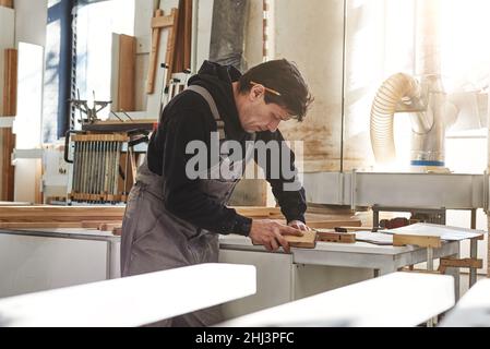 Ritratto di carpentiere lucidare il pannello di legno con carta vetrata in officina. Scatto orizzontale. Vista laterale Foto Stock