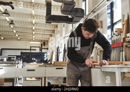 Ritratto di carpentiere in legno uniforme lucidando con carta vetrata in officina. Scatto orizzontale. Vista frontale Foto Stock