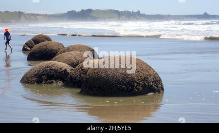 Un ragazzo corre sulla sabbia accanto a grandi pietre (i massi Moeraki) su una spiaggia di South Island NZ. Foto Stock