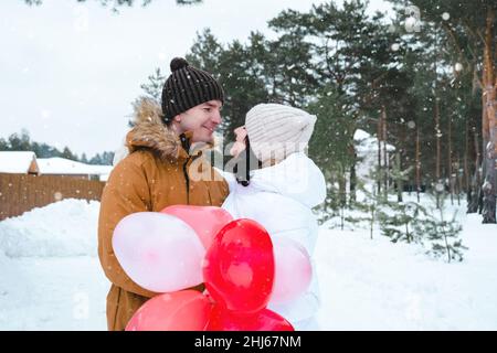 Un uomo e una donna innamorati in una data all'aperto in inverno nella neve con un regalo di palloncini rosa e rosso a forma di cuore. San Valentino, amore Foto Stock