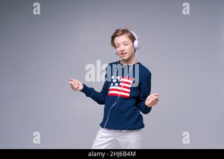 Felice ragazzo divertente indossare cuffie cablate ascoltare musica e mostrare il movimento rock e roll con il viso felice. Studio interno girato isolato su sfondo grigio. In un maglione con bandiera usa Foto Stock