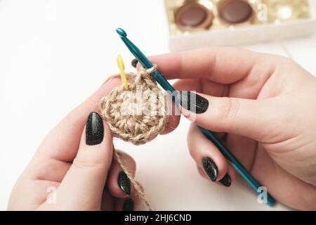 mani di una donna che tiene uncinetto e filo di iuta e frammento di un modello a maglia. maglieria ecologica naturale, montagne russe fatte a mano. artigianato e hobby, arredamento per la casa eco Foto Stock