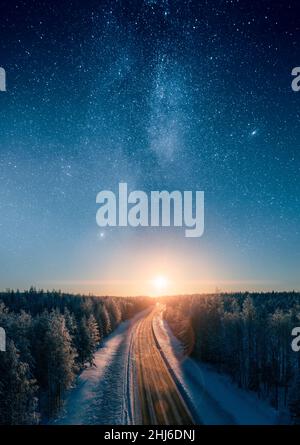 Strada che conduce verso il tramonto e stelle del modo lattiginoso attraverso nevoso foresta boreale. Trasporti e paesaggi epici di nordic. Foto Stock