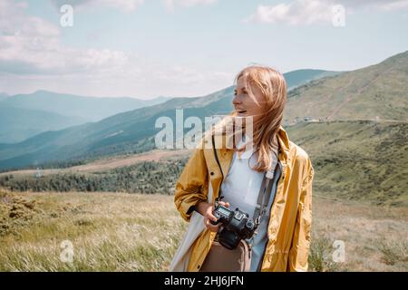 Bella ragazza bionda sorriso turista in giallo impermeabile e con una macchina fotografica guarda le belle colline verdi dei Carpazi di Ucraina su un sole Foto Stock