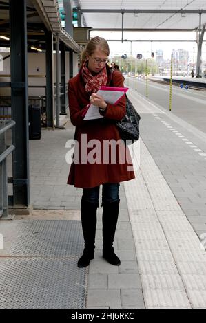 Rotterdam, Paesi Bassi. Street Ritratto di una donna, giovane adulto commuter in attesa per la sua Intercity Train casa legato dopo una giornata di lavoro, nel frattempo leggere una rivista per uccidere il tempo. Foto Stock