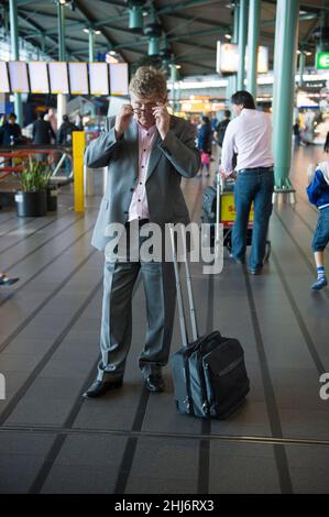 Aeroporto Schiphol, Amsterdam, Paesi Bassi. Uomo caucasico per adulti con valigia e bagaglio a mano/in cabina per una chiamata rapida all'interno del terminal principale dell'aeroporto di Amsterdam. Foto Stock