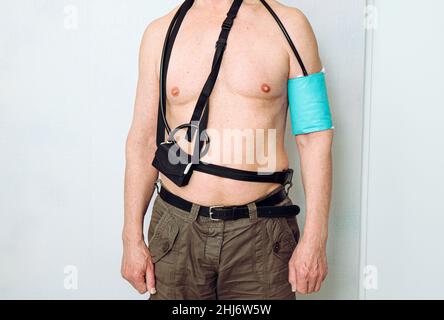 Corpo di un uomo di mezza età che utilizza il monitor della pressione sanguigna ambulatoriale (ABPM) portatile per effettuare le misurazioni durante le normali attività quotidiane a casa. Foto Stock