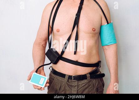 Corpo di un uomo di mezza età che utilizza il monitor della pressione sanguigna ambulatoriale (ABPM) portatile per effettuare le misurazioni durante le normali attività quotidiane a casa. Foto Stock