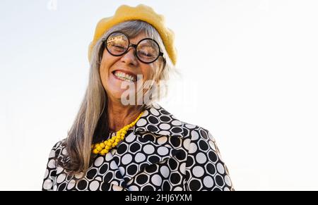 Donna anziana estatica che sorride alla telecamera mentre si trova in piedi all'aperto. Donna anziana felice indossando abiti colorati casual in estate. Tappetino spensierato Foto Stock