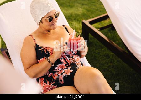 Donna matura che ha un cocktail tiki mentre si rilassa su un lettino. Donna anziana spensierata godendo la sua vacanza estiva in un resort termale di lusso. Senior WO Foto Stock