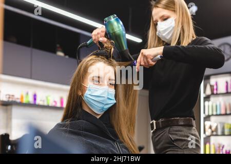 Master donna parrucchiere in una maschera medica asciuga i capelli della ragazza con un asciugacapelli e pettini dopo il lavaggio in un salone di bellezza. Covid-19 pandemia e. Foto Stock