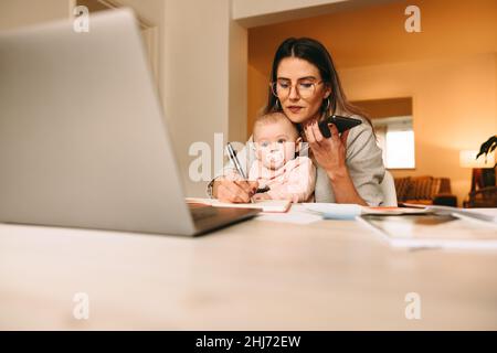 Mamma che lavora prendendo appunti durante una telefonata con i suoi clienti. Mamma multitasking che lavora a un nuovo progetto creativo nel suo ufficio domestico. Interno femmina Foto Stock