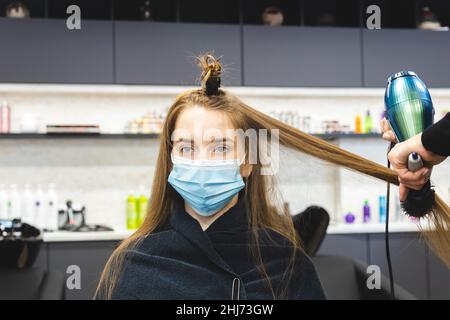 Master donna parrucchiere in una maschera medica asciuga i capelli della ragazza con un asciugacapelli e pettini dopo il lavaggio in un salone di bellezza. Covid-19 pandemia e. Foto Stock