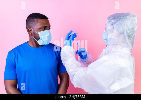 African american man scienziato in PPE suite uniforme mostrando medicina liquido vaccino flacone flacone coronavirus studio parete rosa Foto Stock