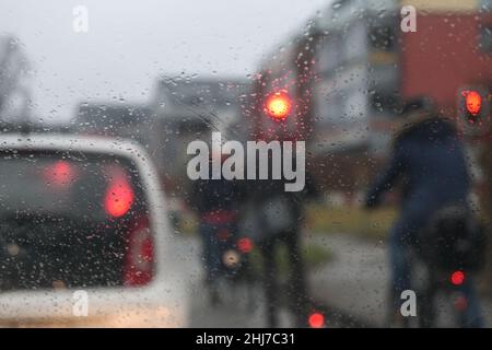 Pioggia sul parabrezza mentre si attende di fronte a un semaforo rosso, ciclisti sfuocati, auto e case all'ora di punta in una giornata piovosa in città Foto Stock
