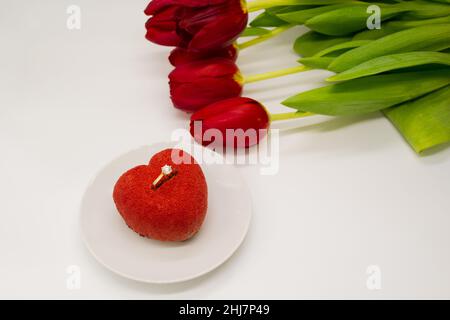 Un bouquet di tulipani rossi accanto ad un piatto con una torta a forma di cuore, in cui un anello d'oro con un diamante. Foto Stock