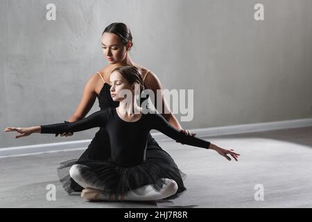 giovane insegnante di balletto che assiste la ragazza seduta con le gambe incrociate durante la lezione Foto Stock