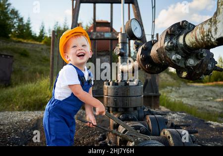 Bambino maschio felice che gioca nel meccanico, tenendo la chiave vicino pipeline del carro di perforazione. Bambino ragazzo in casco da costruzione che studia nuove abilità. Divertimento per l'infanzia all'aperto. Foto Stock