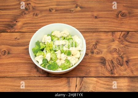 Ciotola bianca riempita di broccoli e fiori di cavolfiore posti su un fondo di legno di quercia colorato Foto Stock