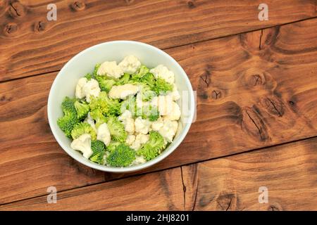 Ciotola bianca riempita di broccoli e fiori di cavolfiore posti su un fondo di legno di quercia colorato Foto Stock