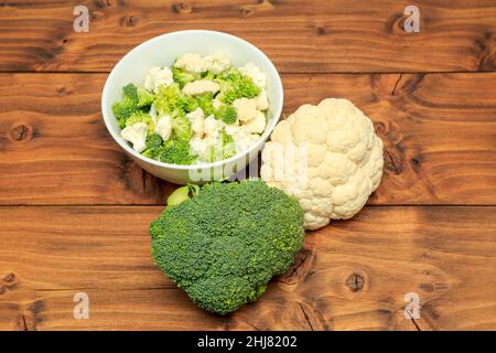 Ciotola bianca riempita con fiori di broccoli e cavolfiore con teste di broccoli e cavolfiore poste su fondo di legno di quercia Foto Stock