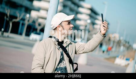 un uomo di mezza età, che indossa abiti casual moderni colorati, sta facendo una videochiamata o prendendo un selfie con il suo smartphone, all'aperto, in uno spazio urbano Foto Stock