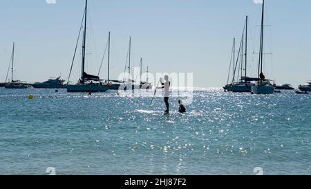 Padre e figlio in piedi su un paddleboard vicino alla spiaggia sull'isola di Formentera in Spagna. Sullo sfondo ormeggiate barche a vela Foto Stock