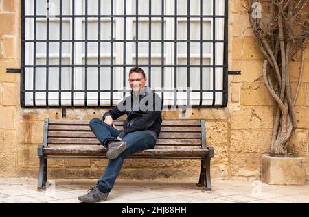 Ritratto di un uomo bianco di 42 anni fiducioso e rilassato, seduto su una panchina con uno sfondo storico facde Foto Stock