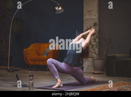 La giovane donna esegue lo yoga stretching facendo Anjaneyasana o bassa affondo posa in moderna casa interna. Donna che pratica yoga a casa. Concetto di asana a casa. Concetto di corpo sano. Concetto di meditazione. Foto Stock