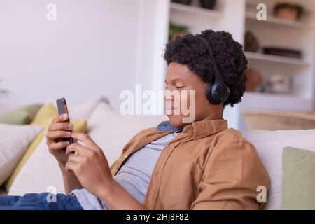 Sorridente giovane afroamericano che indossa le cuffie mentre ascolta la musica sullo smartphone a casa. Inalterato, stile di vita, attività per il tempo libero, wireless t Foto Stock