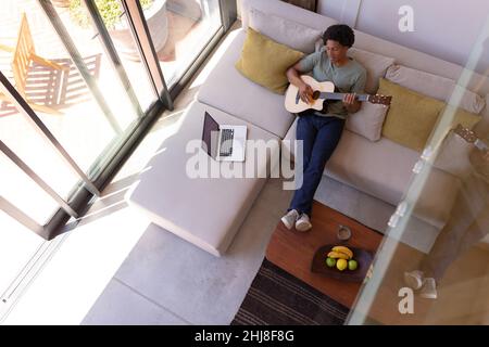 Giovane afroamericano che suona la chitarra mentre si siede con il portatile e i piedi sul tavolo a casa. Inalterato, musica, laptop, lifestyle, cibo, bere, le Foto Stock
