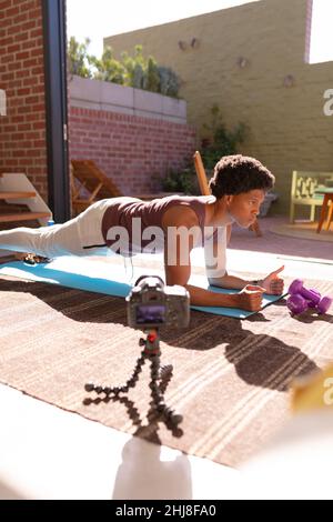 afroamericano giovane uomo che fa push-aumenta vlog a casa. Inalterato, fitness, vlogging, stretching, macchina fotografica, dumbbell, influencer e stile di vita attivo c Foto Stock