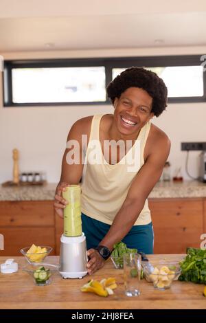 Ritratto del giovane afroamericano sorridente che prepara il frullato in juicer a casa. Inalterato, influencer, cibo sano, biologico e sano stile di vita Foto Stock