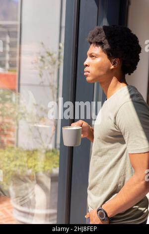 Vista laterale del giovane afro-americano con una tazza di caffè in piedi dalla finestra a casa. Inalterato, stile di vita, bevanda, contemplazione, acconciatura afro Foto Stock
