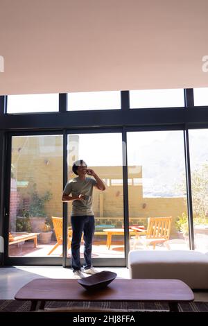 Felice afroamericano giovane che parla al telefono mentre si sta in piedi con una tazza di caffè dalla finestra. Inalterato, stile di vita, attività di svago, drink, commun Foto Stock