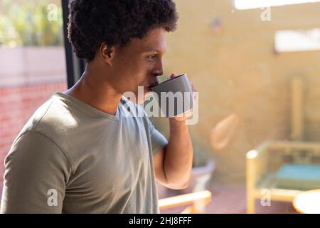 Primo piano del giovane afro-americano che beve il caffè mentre si alza accanto alla finestra a casa. Inalterato, stile di vita, attività di svago, drink, rinfresco e Foto Stock
