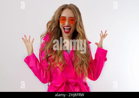 Primo piano gridando ragazza felice in occhiali da sole rossi, cappotto rosa isolato su sfondo bianco. Eccitato, shock e sorpresa giovane donna in abito alla moda. Fa Foto Stock