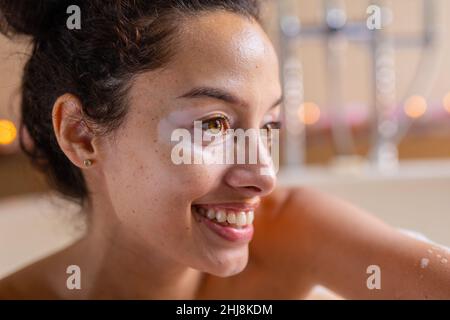 Primo piano di sorridente ragazza biraciale bella guardando via nella vasca da bagno al centro benessere. Inalterato, spa, cura del corpo, stile di vita, vitiligo e benessere. Foto Stock