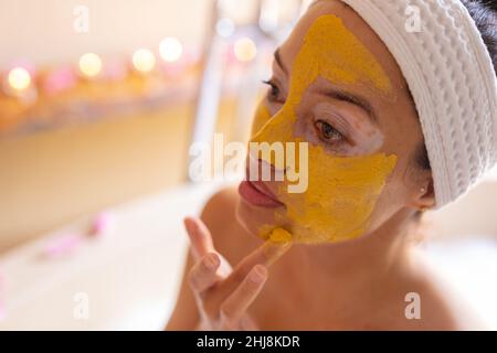 Primo piano della giovane donna biraciale che applica la maschera facciale gialla alla spa. Inalterato, spa, cura del corpo, stile di vita, vitiligo e benessere. Foto Stock