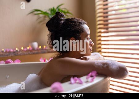 Vista laterale della giovane donna biraciale pensata che guarda attraverso le tende della finestra mentre si appoggia sulla vasca da bagno. Inalterato, spa, cura del corpo, stile di vita, solitudine, Foto Stock